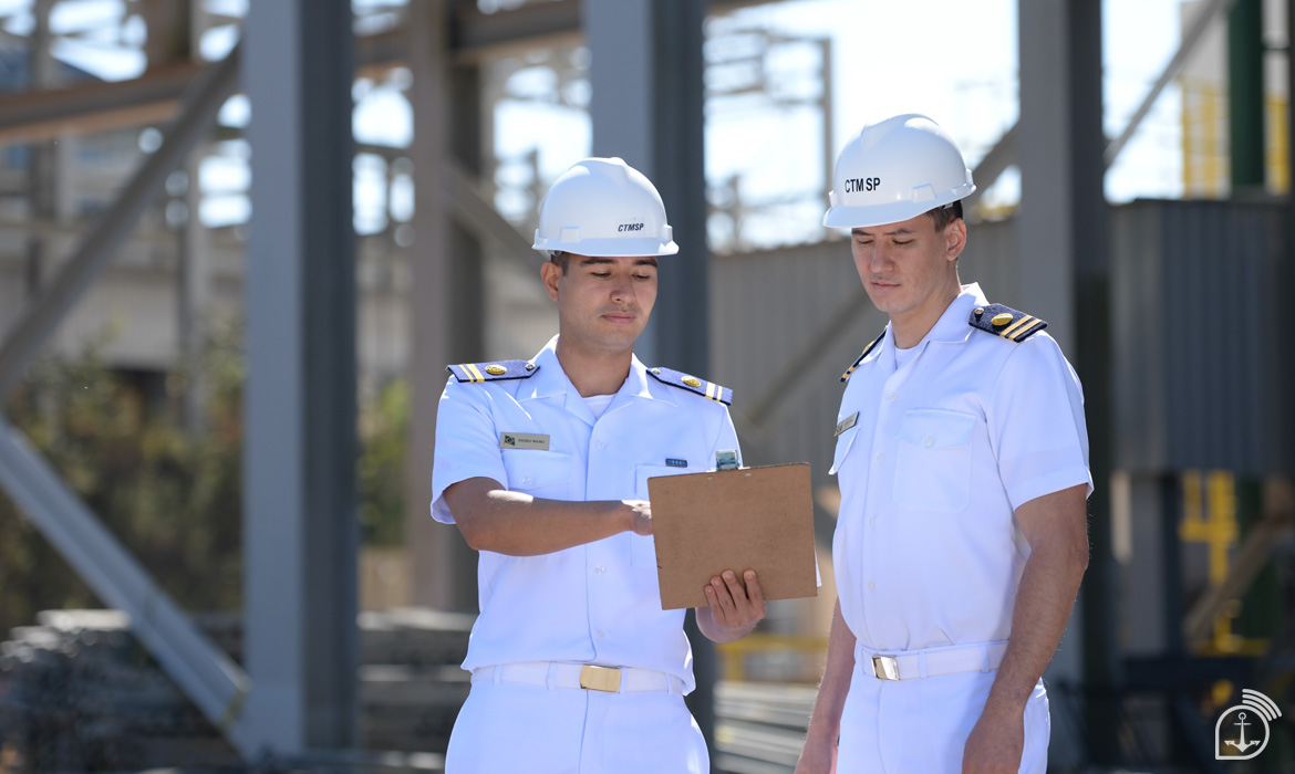 Dois oficiais da Marinha do Brasil, em uniformes brancos e capacetes, analisam documentos em uma instalação industrial. Imagem: Marinha do Brasil