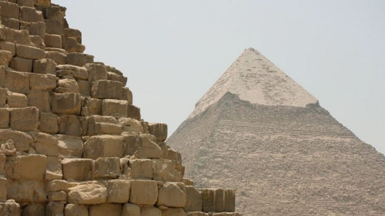 Fotografia real capturada no complexo de Gizé, no Egito, mostra em primeiro plano os enormes blocos de calcário da Pirâmide de Miquerinos. Ao fundo, destaca-se a imponente Grande Pirâmide de Quéops, com parte de seu revestimento original ainda visível no topo. A imagem evidencia a escala monumental e a precisão da engenharia egípcia antiga.