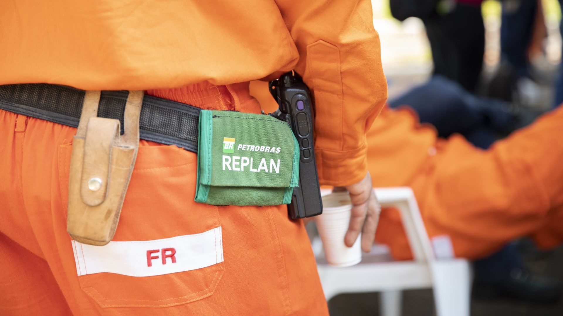 Trabalhador da Petrobras na Replan em uniforme de segurança laranja, com equipamentos e bolsa identificada, durante pausa em área externa da refinaria.