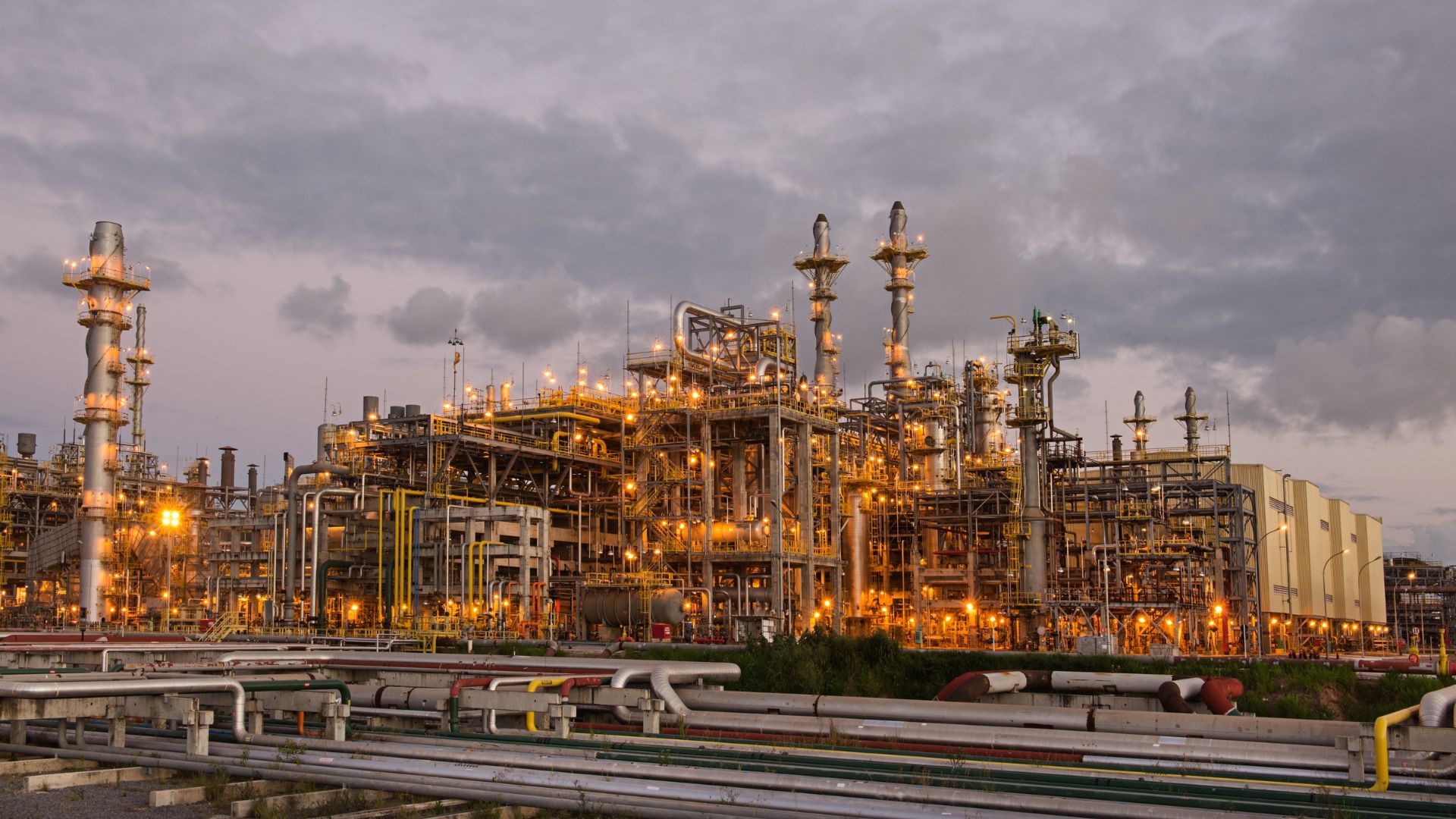 Refinaria de petróleo iluminada ao entardecer, com torres e tubulações em destaque sob céu nublado.