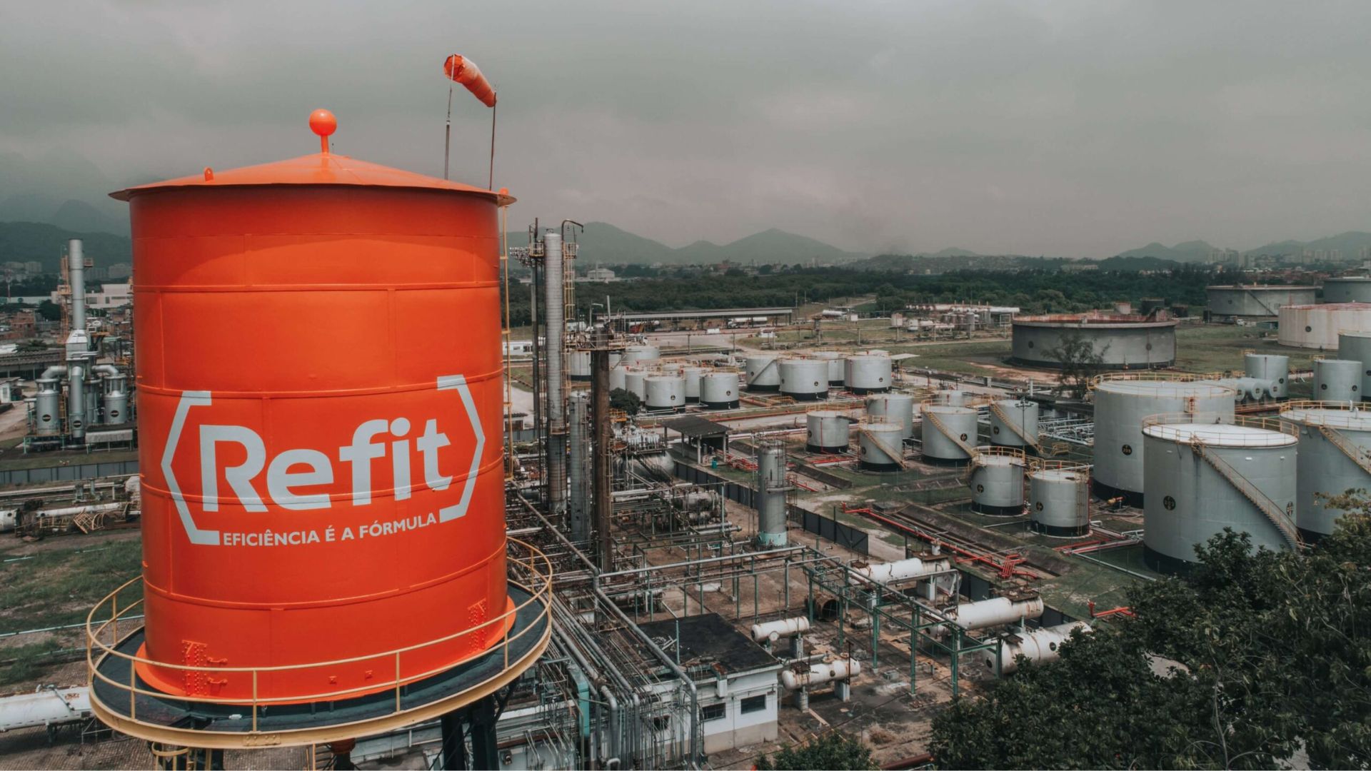 Tanque laranja elevado com a marca Refit em uma refinaria industrial, com infraestrutura de armazenamento e montanhas ao fundo.