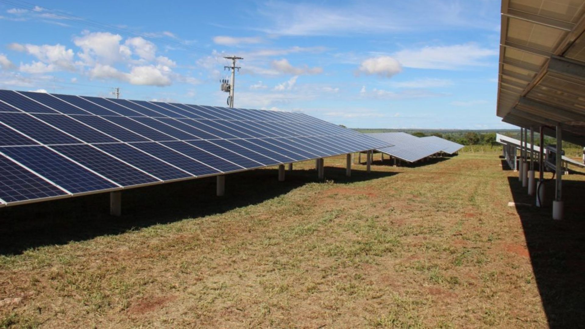 Painéis solares instalados em campo gramado, alinhados em fileiras, com céu limpo ao fundo e conexão à rede elétrica visível