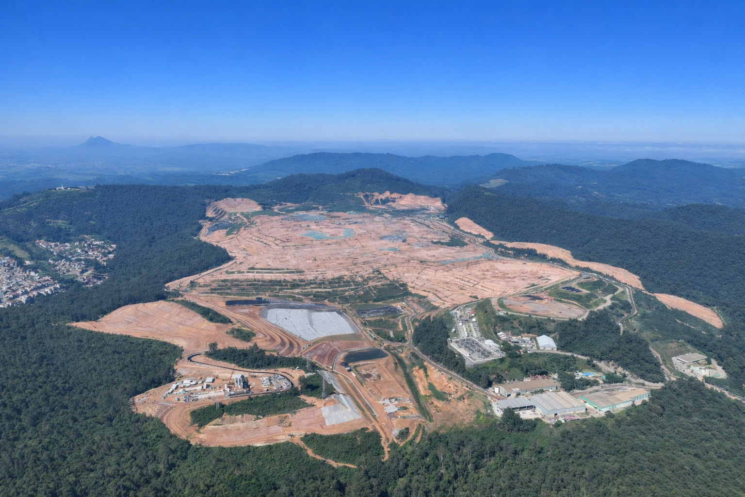 Vista aérea de grande área de construção em meio à mata, com iluminação de meio-dia e céu azul limpo.