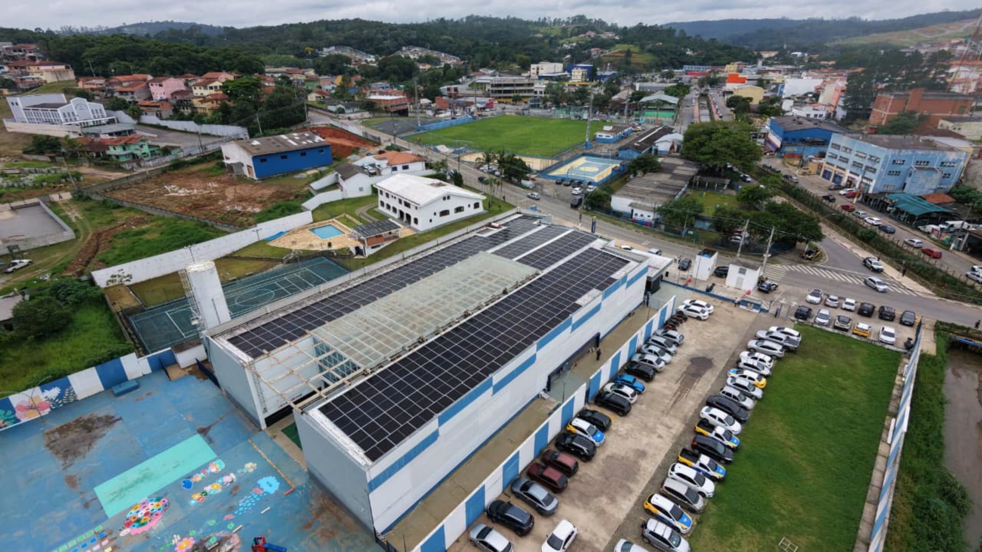 Vista aérea de prédio público com painéis solares no telhado em Cotia, mostrando estacionamento ao lado e quadras esportivas próximas, destacando uso de energia solar.