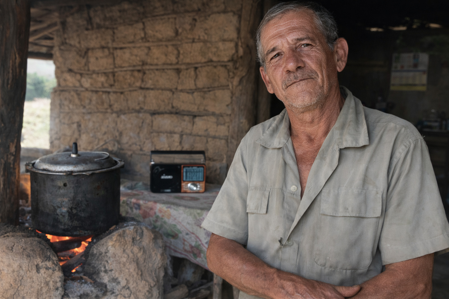 Trabalhador rural em sítio isolado acendendo fogão a lenha com rádio portátil ao lado.